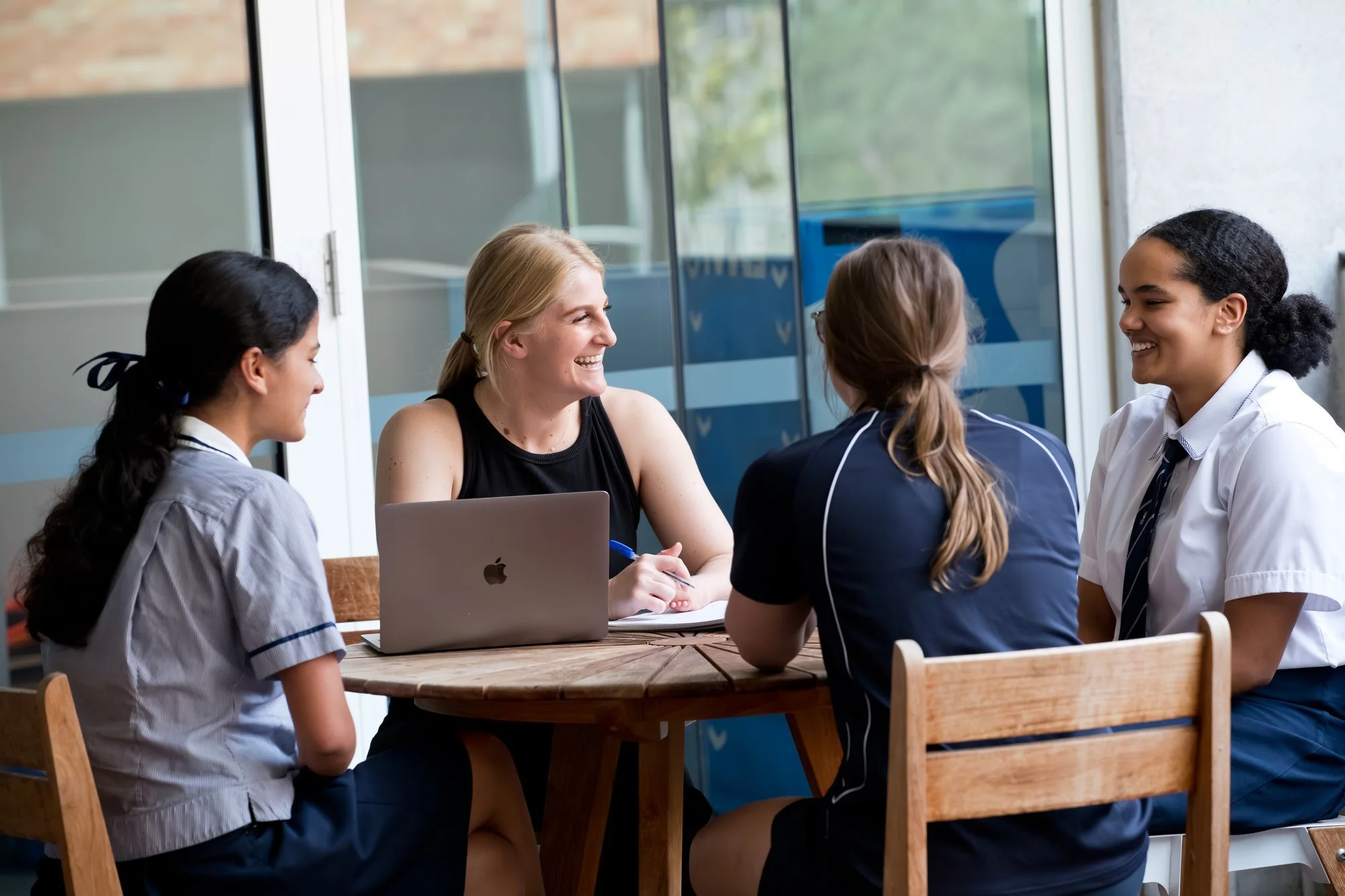 A group of four women, three wearing school uniforms, and one in casual attire, sitting around a wooden table. The woman in casual attire has blonde hair and is smiling while talking to the women in uniforms. A laptop is on the table. Large glass win