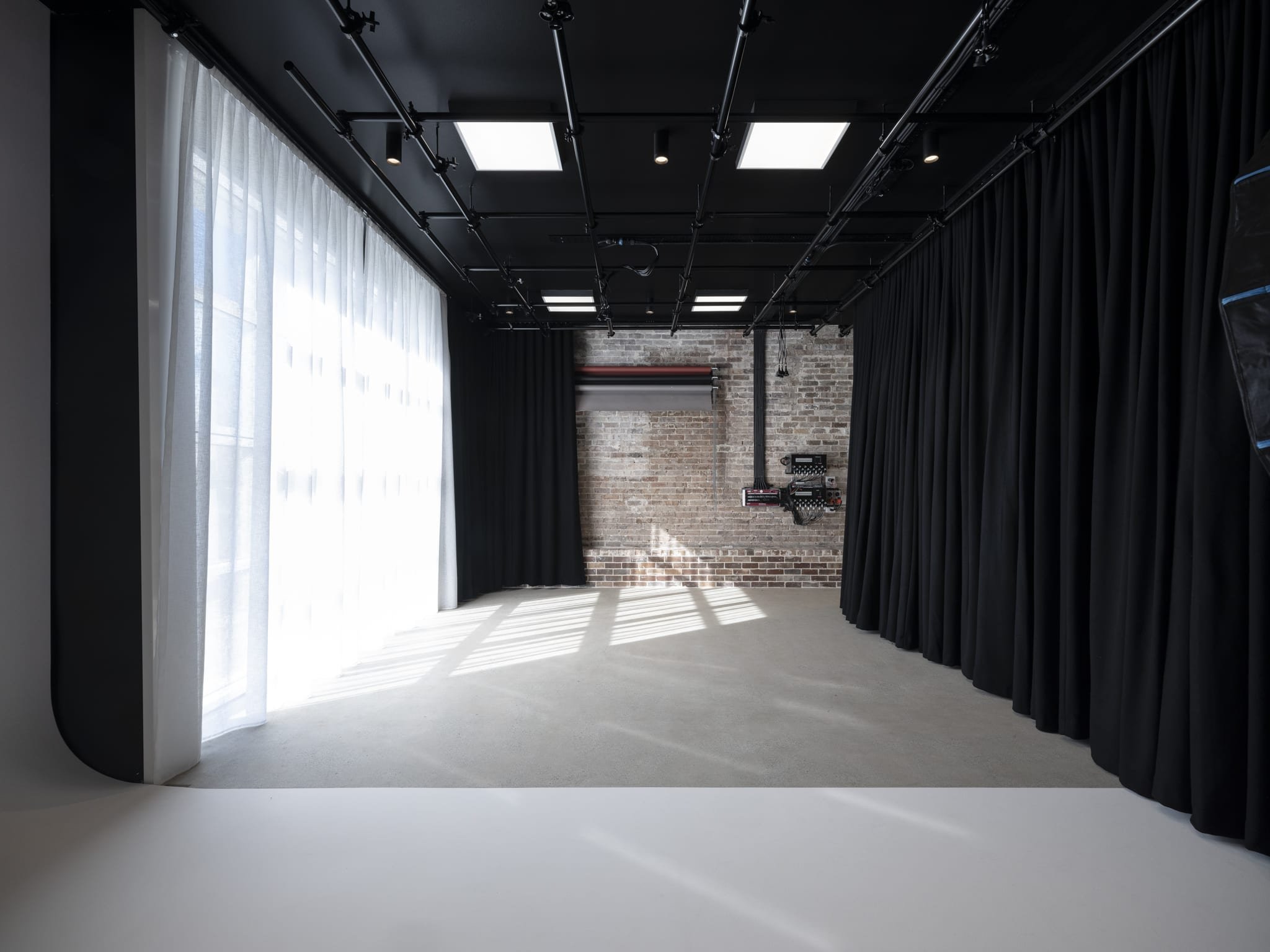 Empty photography or theater studio with black curtains, white sheer curtains on the left, brick wall in the background, studio lighting panels on the ceiling, and some equipment on the brick wall.