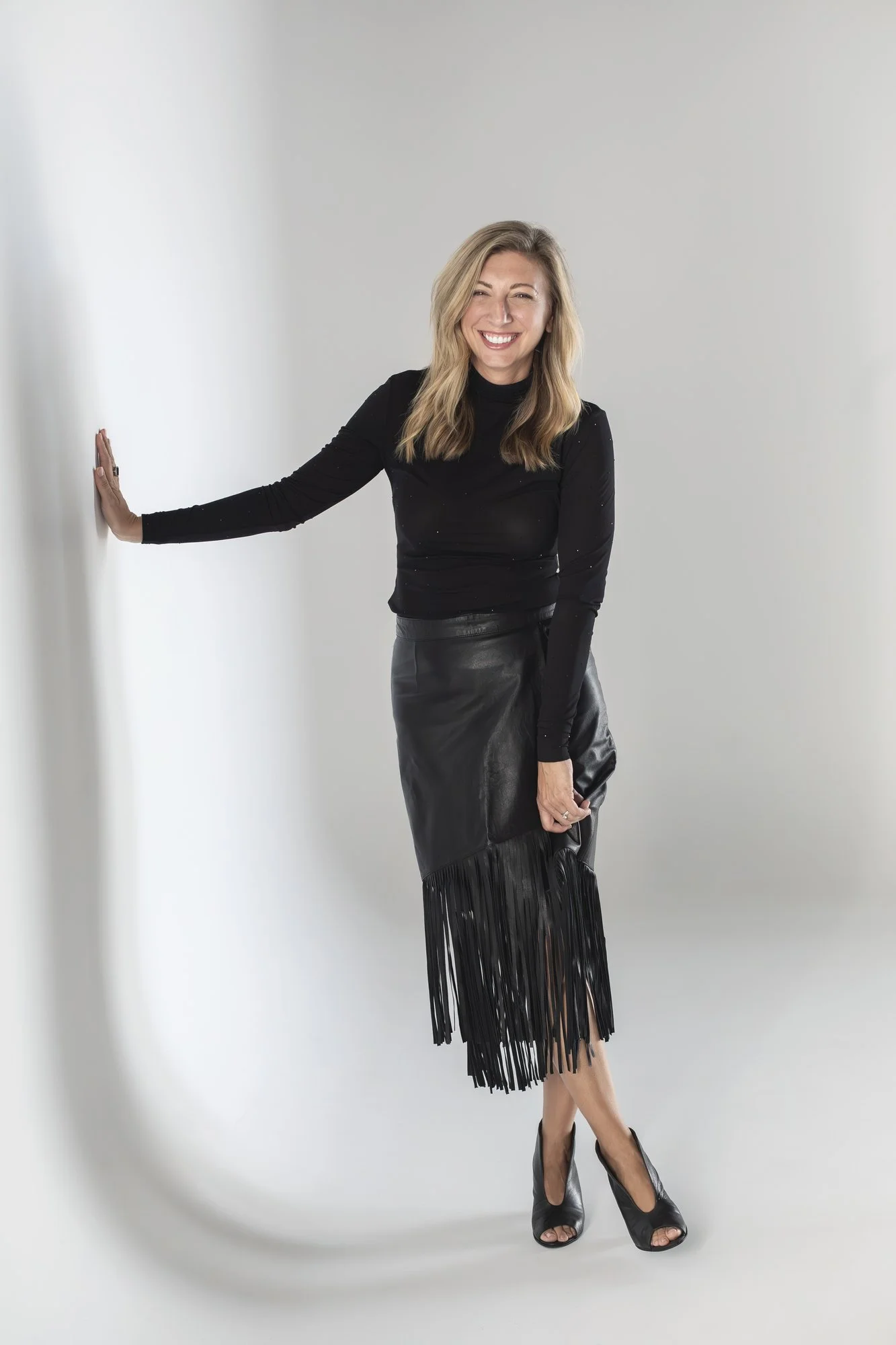 A woman with blonde hair wearing a black long sleeve top, a black leather fringed skirt, and black shoes, standing against a plain gray background with her hand pressed against the wall, smiling and looking at the camera.