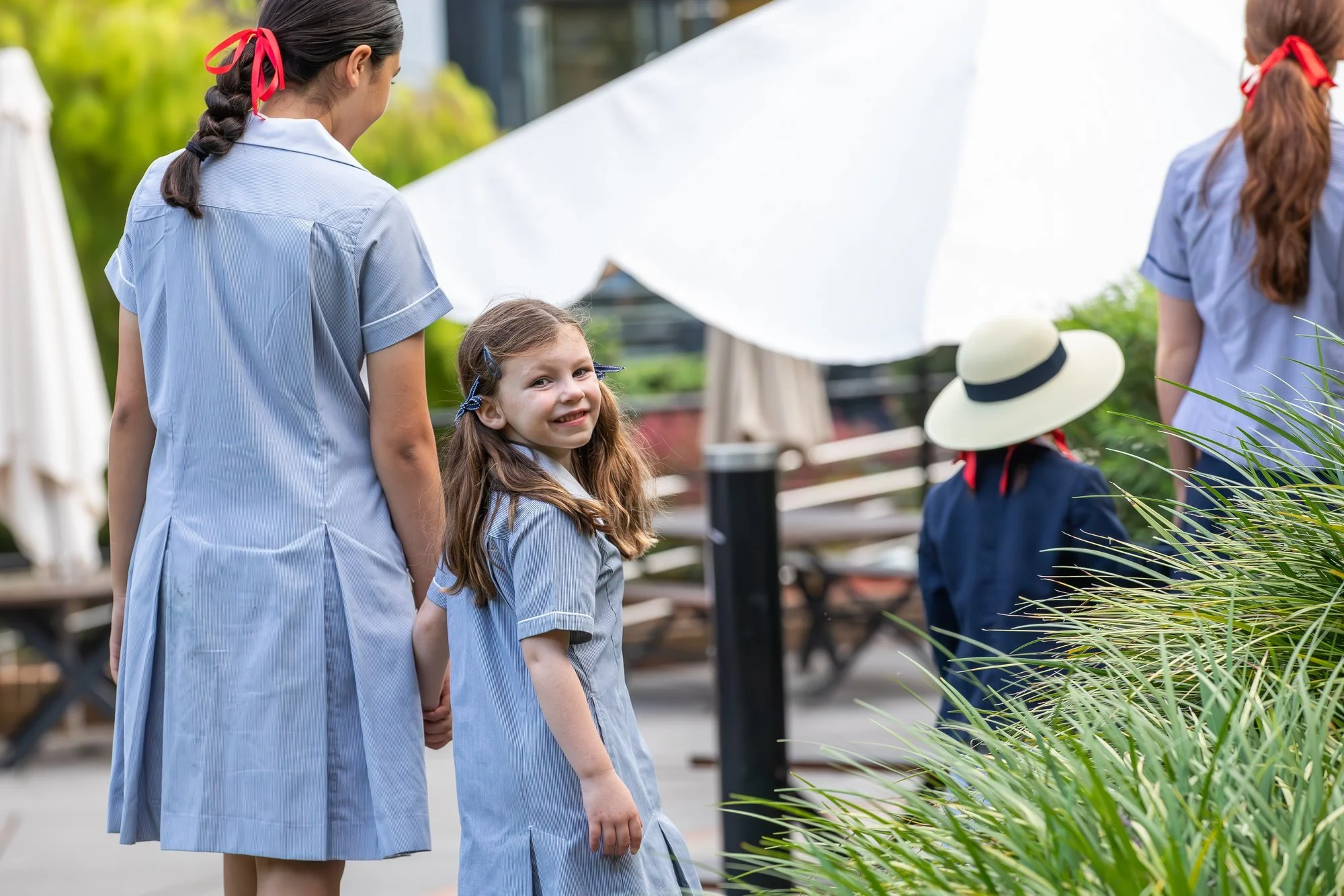 Group of children with school uniforms holding hands outdoors in a garden or park, smiling, with greenery and plants in the background.