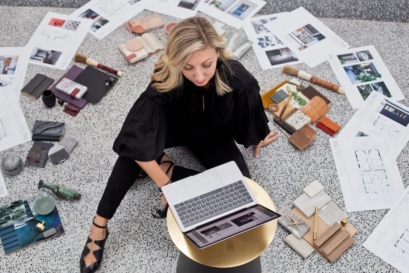 A woman sitting on a terrazzo floor surrounded by interior design materials, color swatches, fabric samples, paint chips, blueprints, and design tools, working on a laptop.
