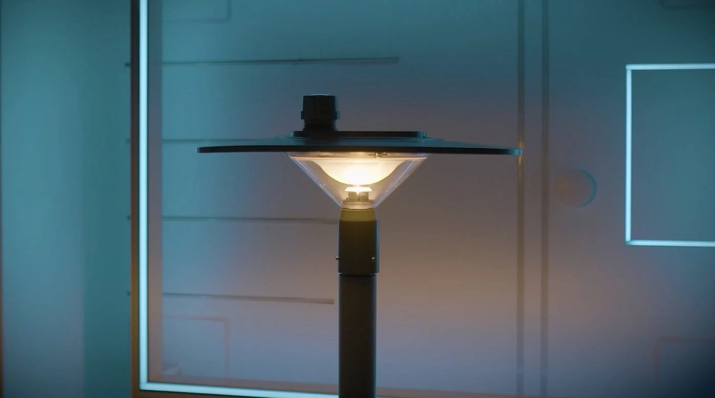 Close-up of a modern outdoor lamp with a glowing bulb, mounted on a pole, against a sleek white building with illuminated trim.