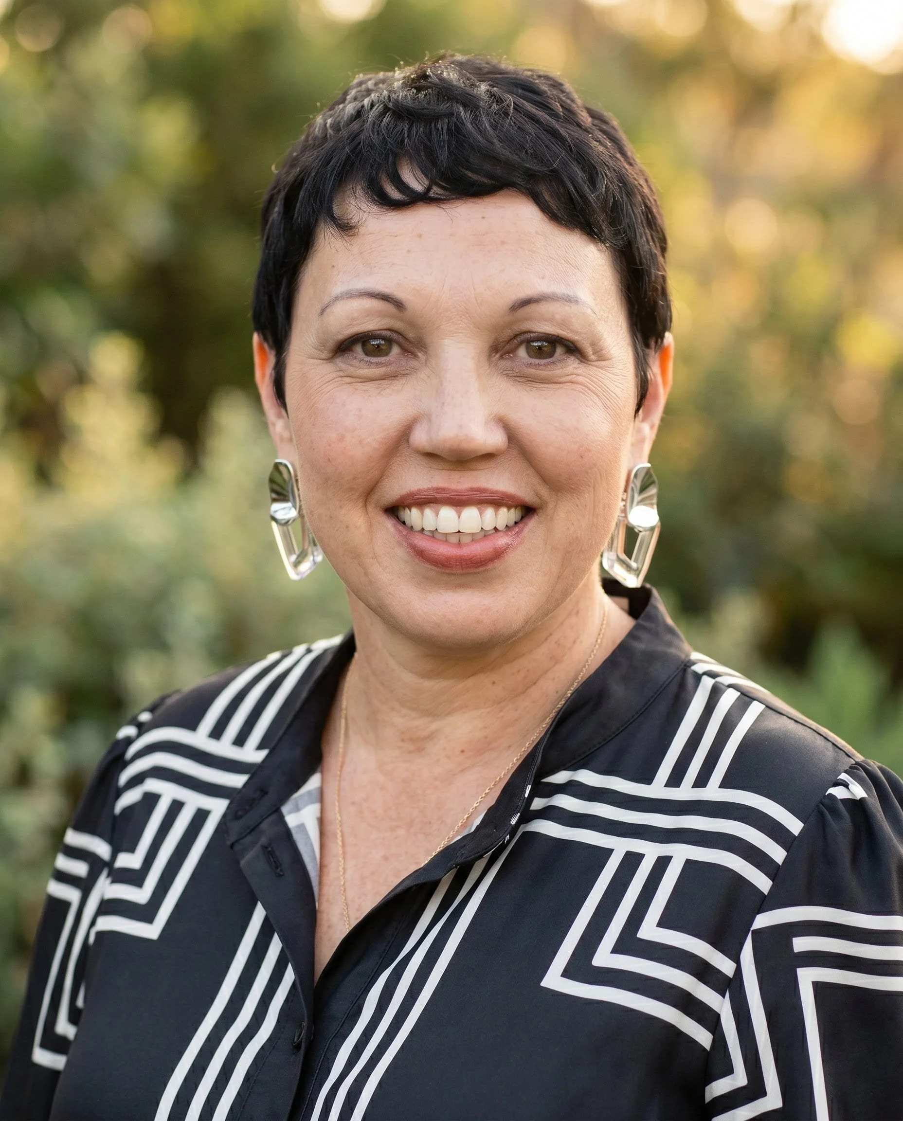 A smiling woman with short black hair and earrings, outdoors with a blurred background of trees and sunlight.