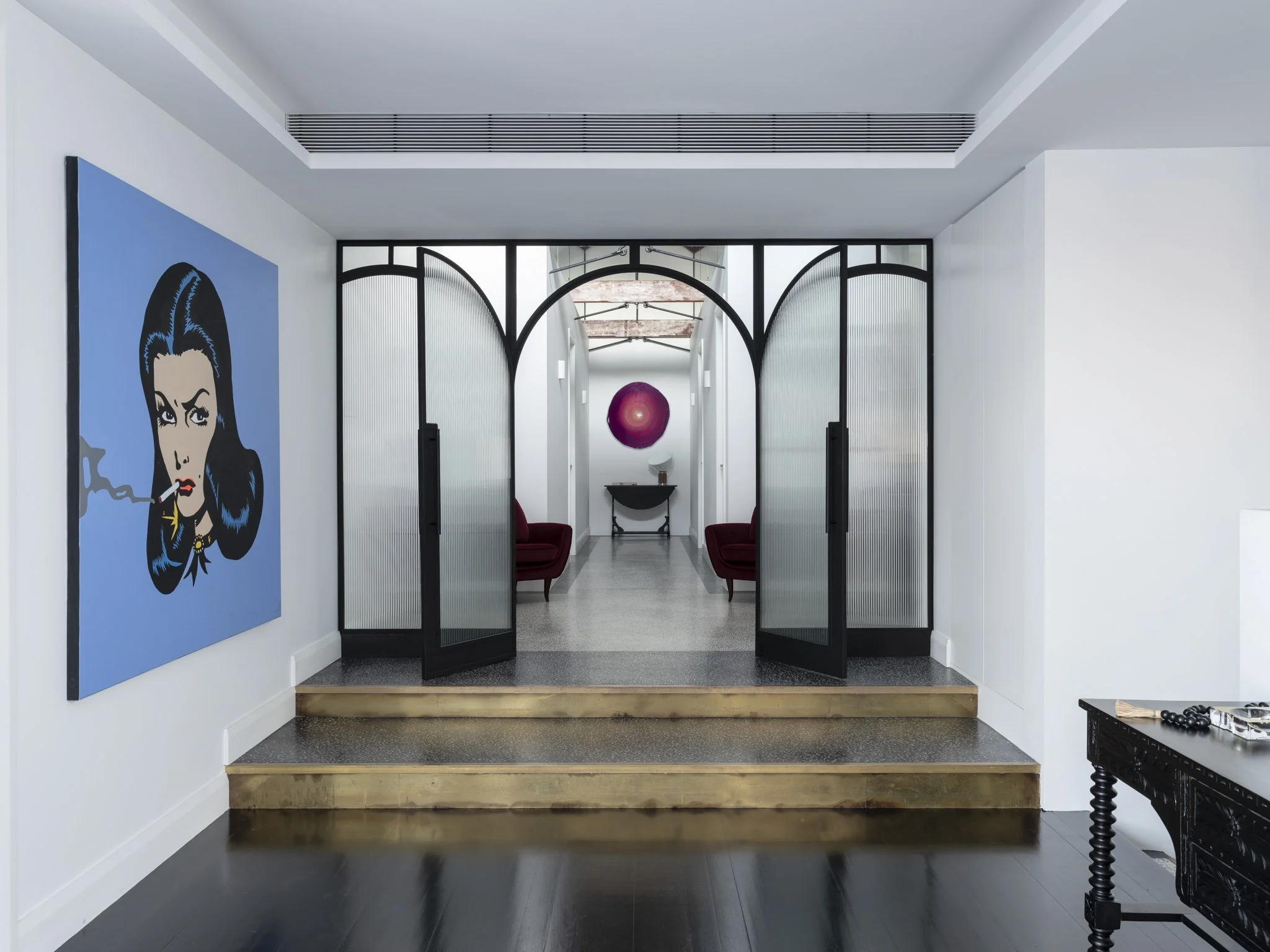Interior hallway with black arched metal sliding doors leading to a sitting area with red chairs, wall art featuring a comic-style woman smoking, and a large pink decorative sphere hanging in the background.