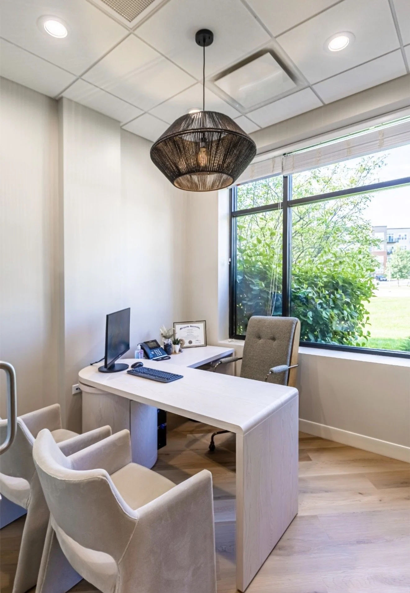 An office with a white desk, computer monitor, keyboard, mouse, telephone, framed certificate, potted plant, and beige chairs. Large window with greenery outside and ceiling light fixtures above.