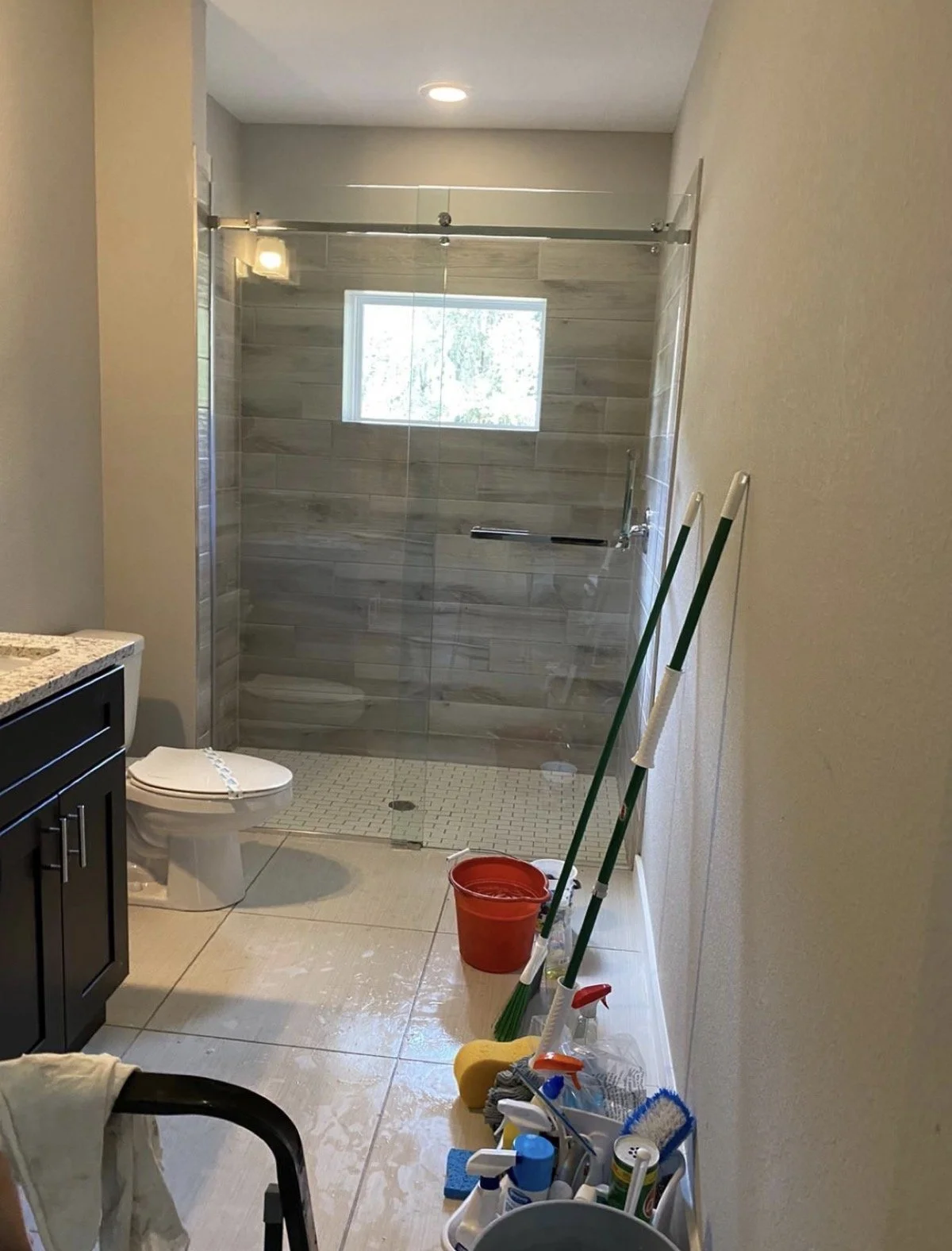 Bathroom with glass shower enclosure, toilet, and cleaning supplies on the floor, including mops, a bucket, and cleaning tools.