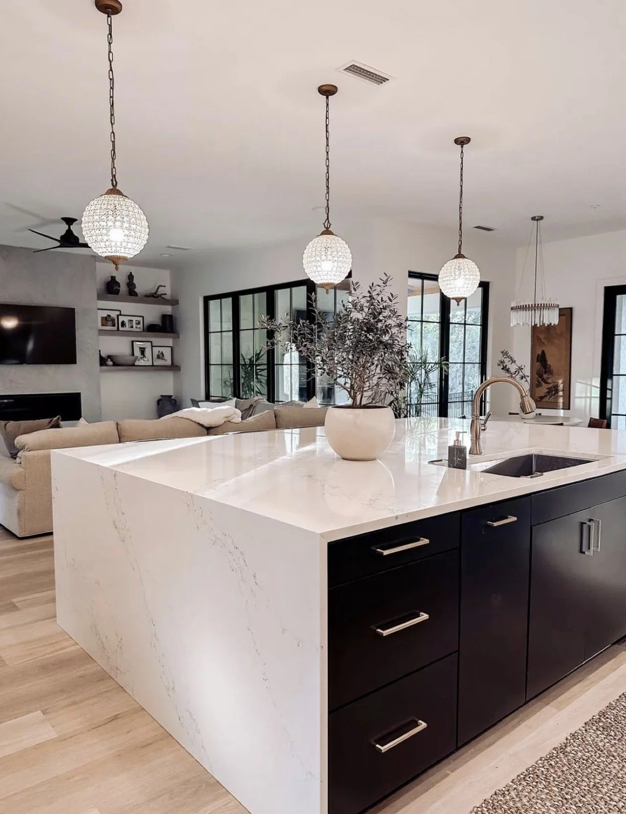 Modern open-concept living room and kitchen with white marble island, black cabinetry, pendant lights, large window with black framing, beige sofa, wall-mounted TV, and decorative shelves.