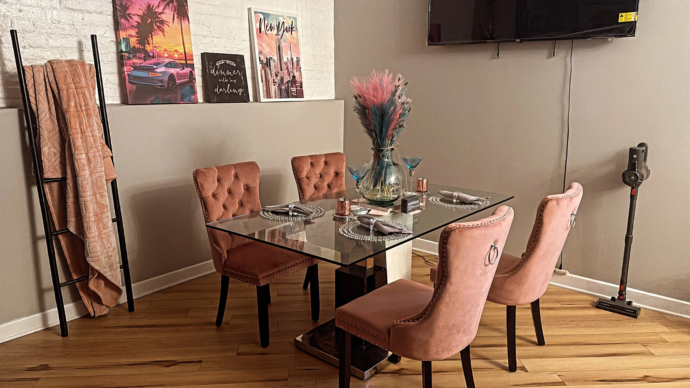 A dining room with a glass table set for four, surrounded by pink velvet chairs. A vase with pink and blue feathers and two blue cocktail glasses are on the table. A wall-mounted TV and a white brick wall with three framed pictures, including one of New York City, are in the background. A black coat rack draped with peach-colored fabrics stands against the wall.