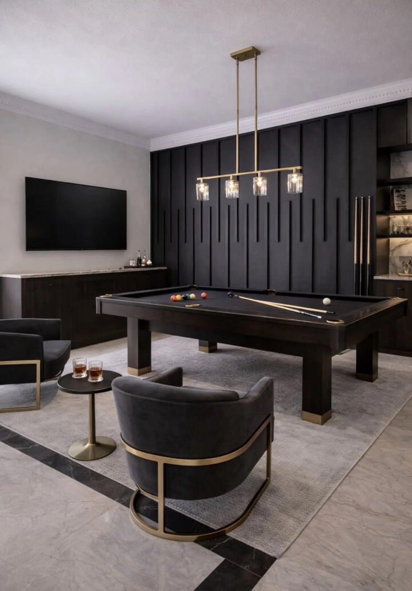 Modern basement game room with a black pool table, gray armchairs, a small round side table with drinks, a wall mounted TV, and a black accent wall with gold accents and a contemporary chandelier.