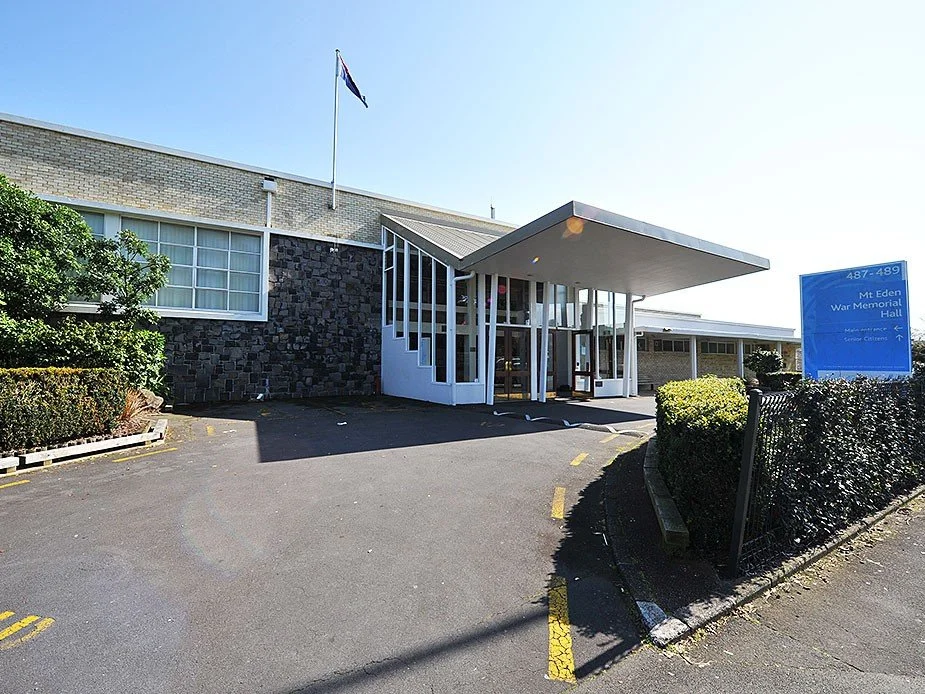 The entrance of Mt. Eden War Memorial Hall with a parking lot in front, nearby bushes, and a blue sign indicating the hall's location.