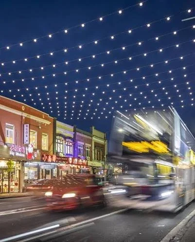 Street scene at night with a double-decker bus and cars, festive string lights overhead, and colorful buildings with shops.