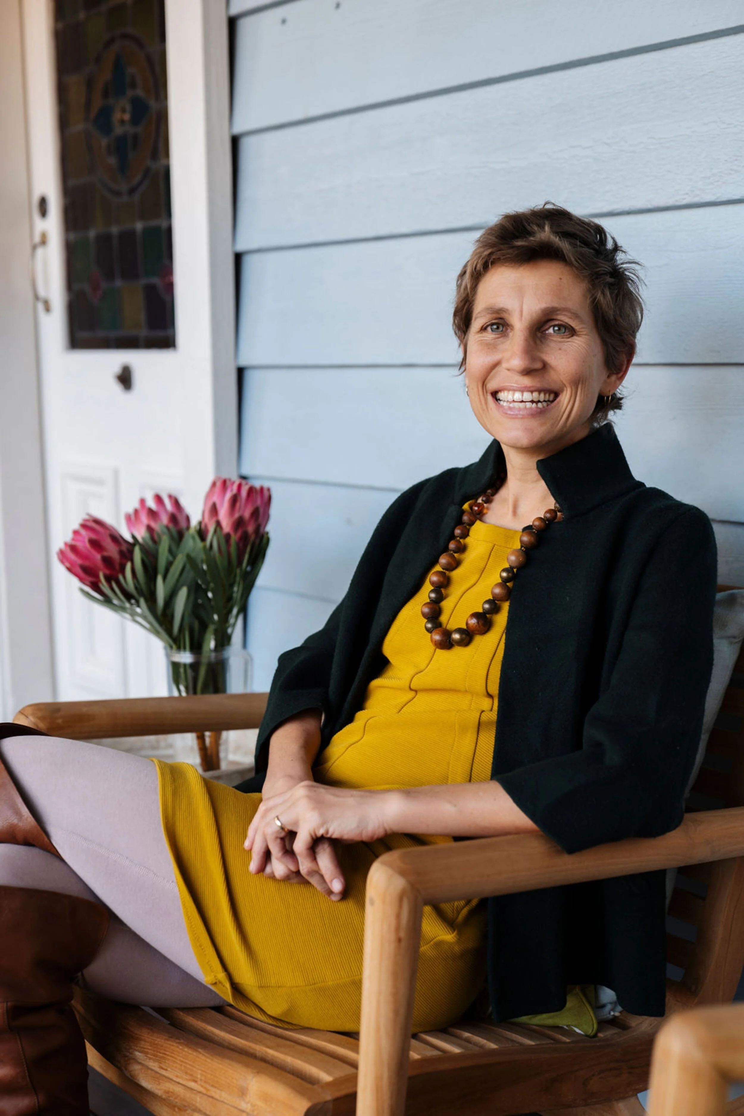 Julia Jones wearing a green blazer, yellow dress, and beaded necklace sitting on a wooden chair outdoors next to a table with a vase of pink flowers, smiling in front of a blue weatherboard house.