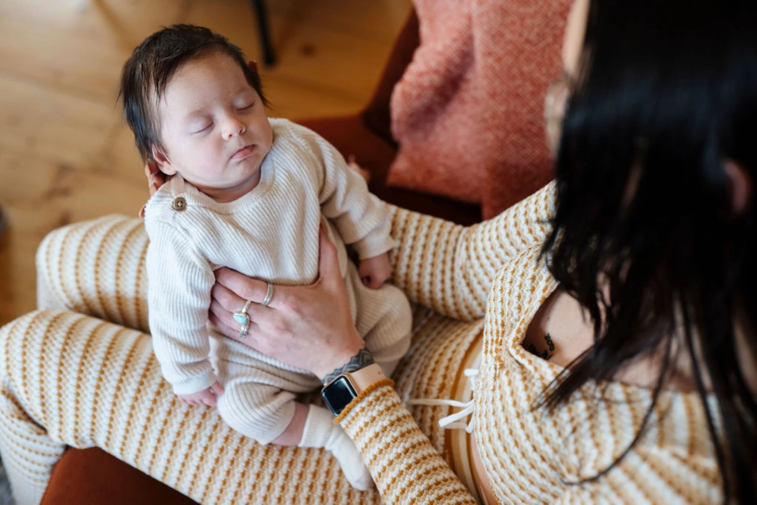 A woman holding a sleeping baby in her arms, both wearing matching beige and brown striped outfits, inside a cozy room with wooden furniture and a red upholstered chair.