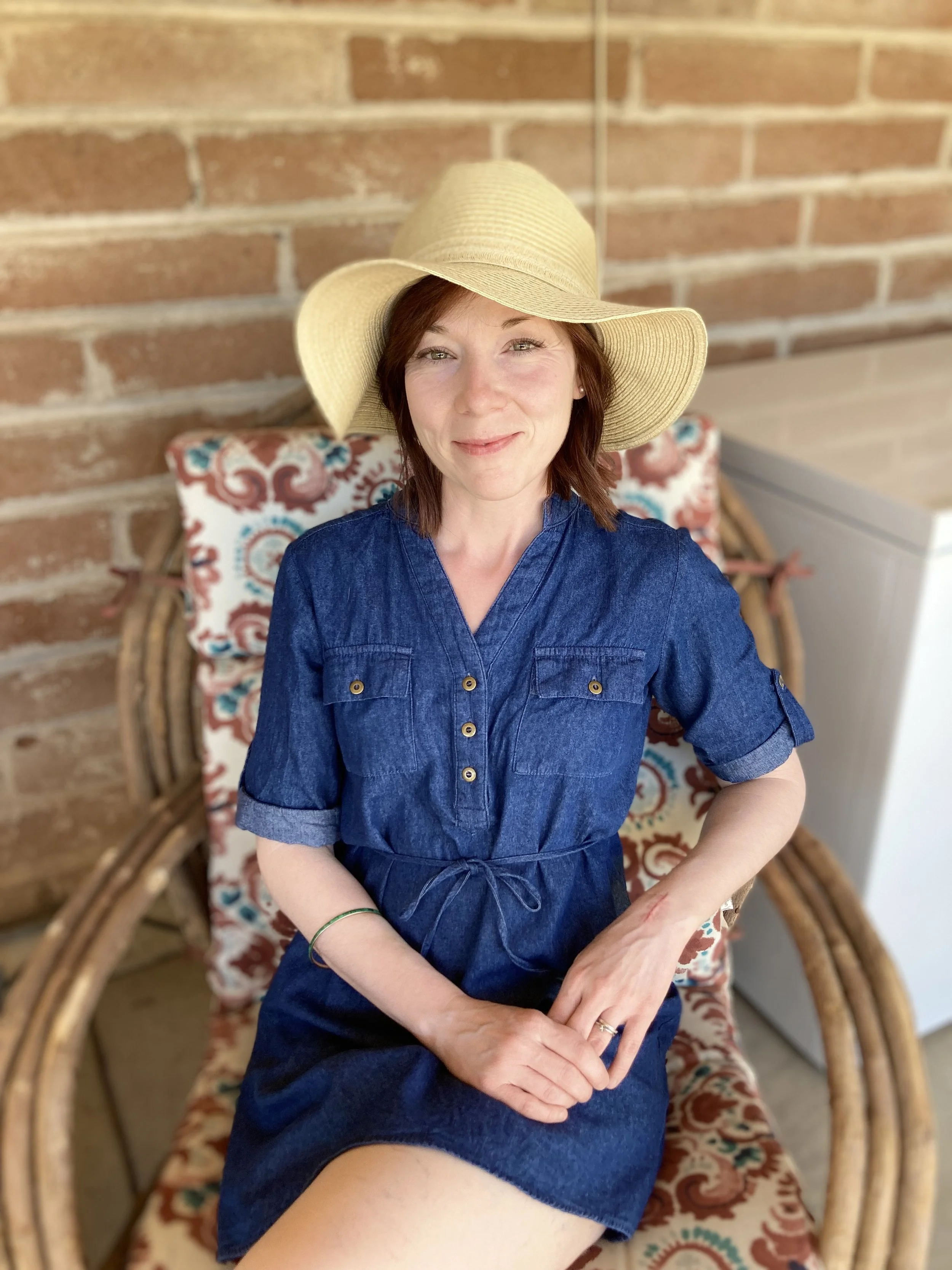 A woman with short brown hair, wearing a wide-brimmed straw hat and a blue denim dress, sitting on a decorative wicker chair with a patterned cushion, in front of a brick wall.