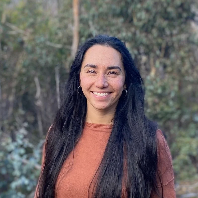 A woman with long dark hair, wearing hoop earrings and an orange shirt, is smiling outdoors with blurred trees and foliage in the background.