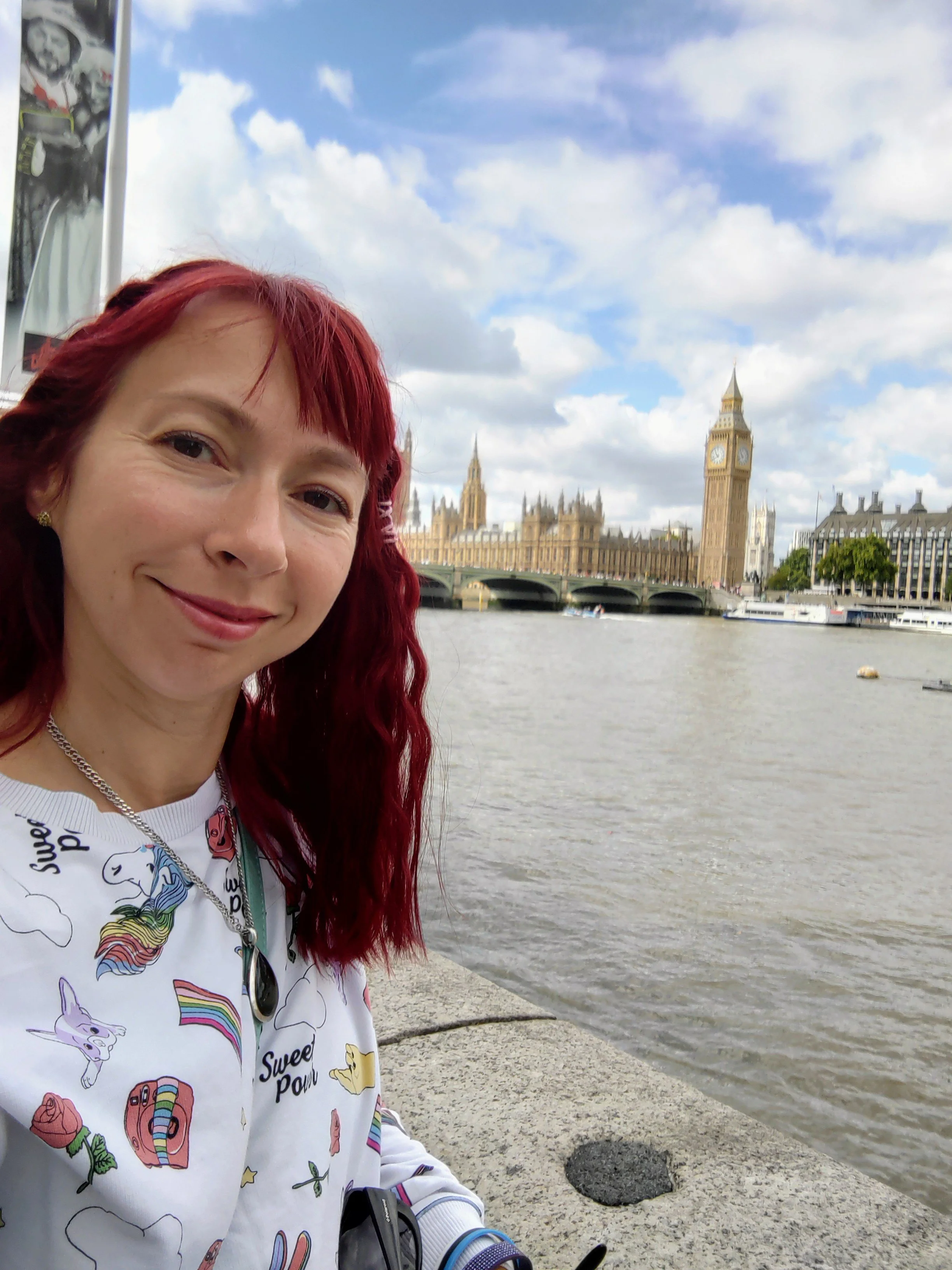 A woman with red hair smiling in front of the London skyline, including Big Ben and the Houses of Parliament, by the river under a partly cloudy sky.