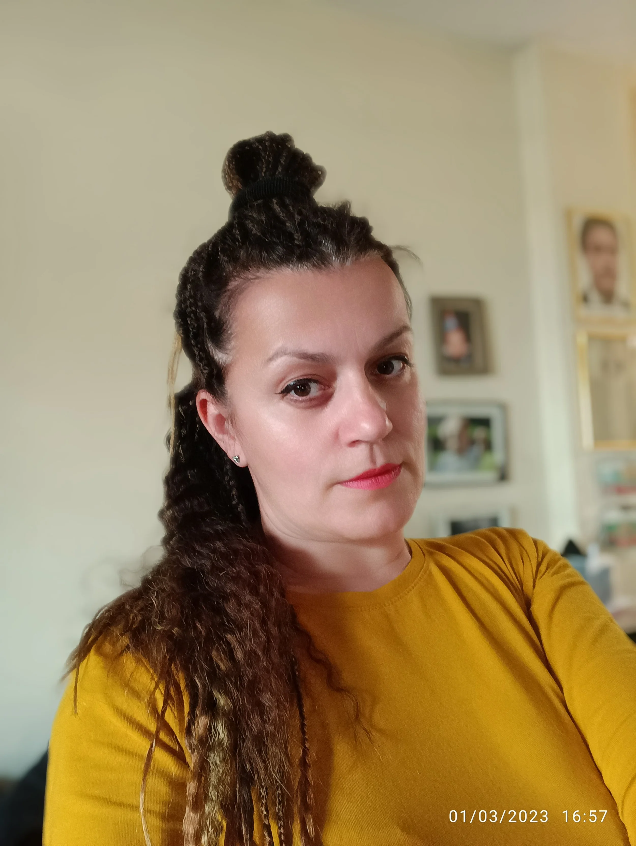 Close-up of a woman with curly brown hair styled in a bun, wearing a mustard yellow shirt, and standing in a room with framed pictures on the wall.