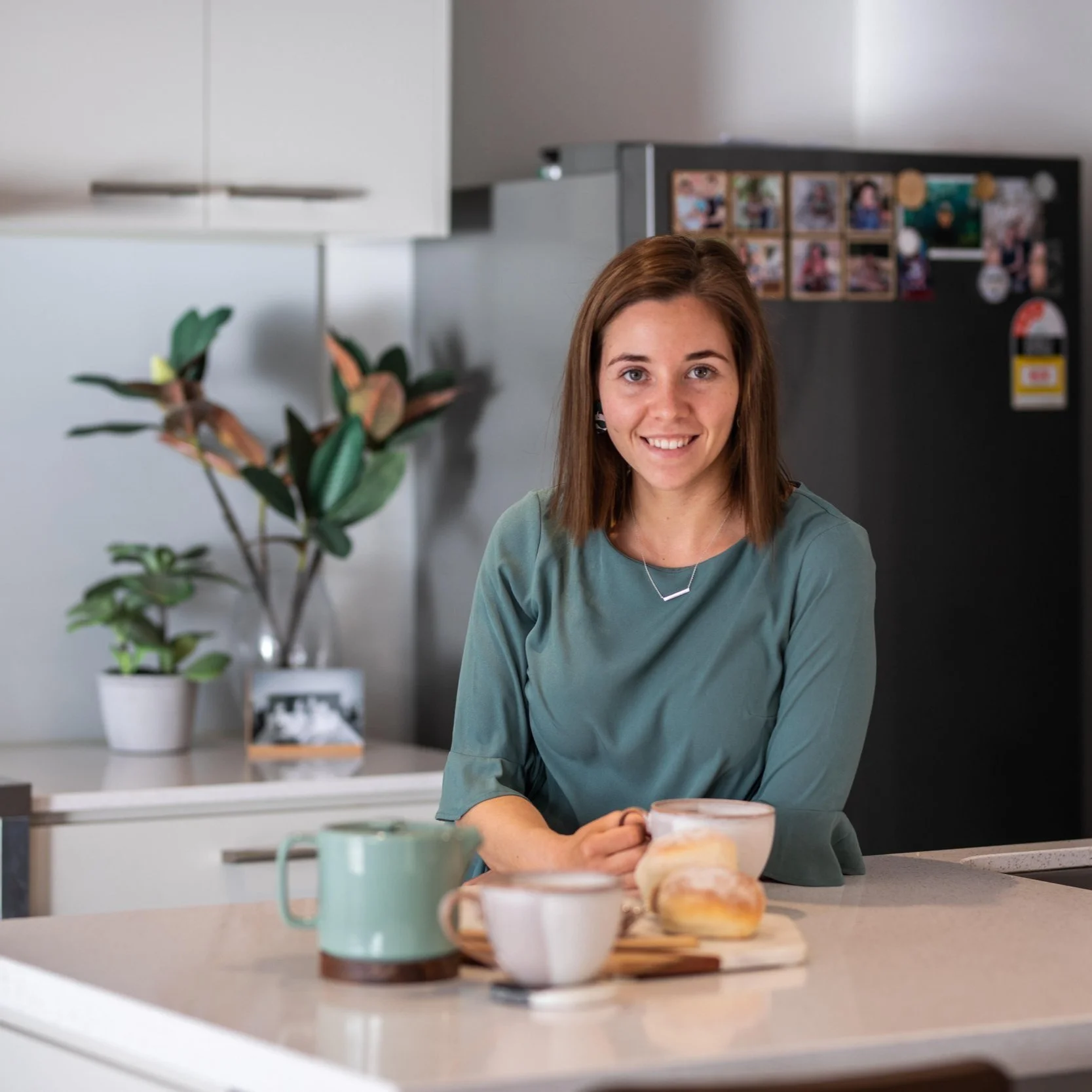 A young woman with shoulder-length brown hair smiling while sitting at a kitchen counter, holding a mug, with coffee and pastries in front of her. The kitchen includes a potted plant, a black refrigerator adorned with photos, and white cabinets.