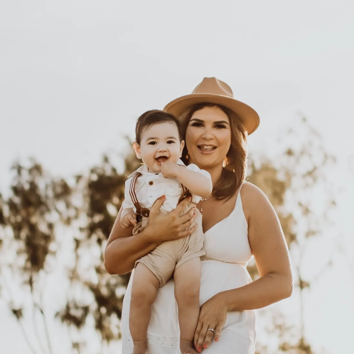 A woman wearing a beige hat and white dress holding a smiling toddler in a white shirt with suspenders outdoors.