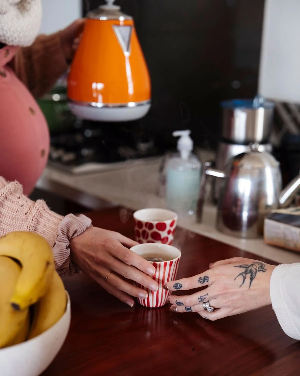 Postpartum professional handing a mother a mug of tea at a kitchen counter, with bananas in a bowl in the foreground.