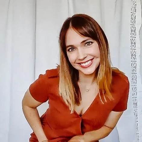 A young woman with shoulder-length light brown hair, wearing a reddish-brown top, smiling and leaning slightly forward against a white curtain background.