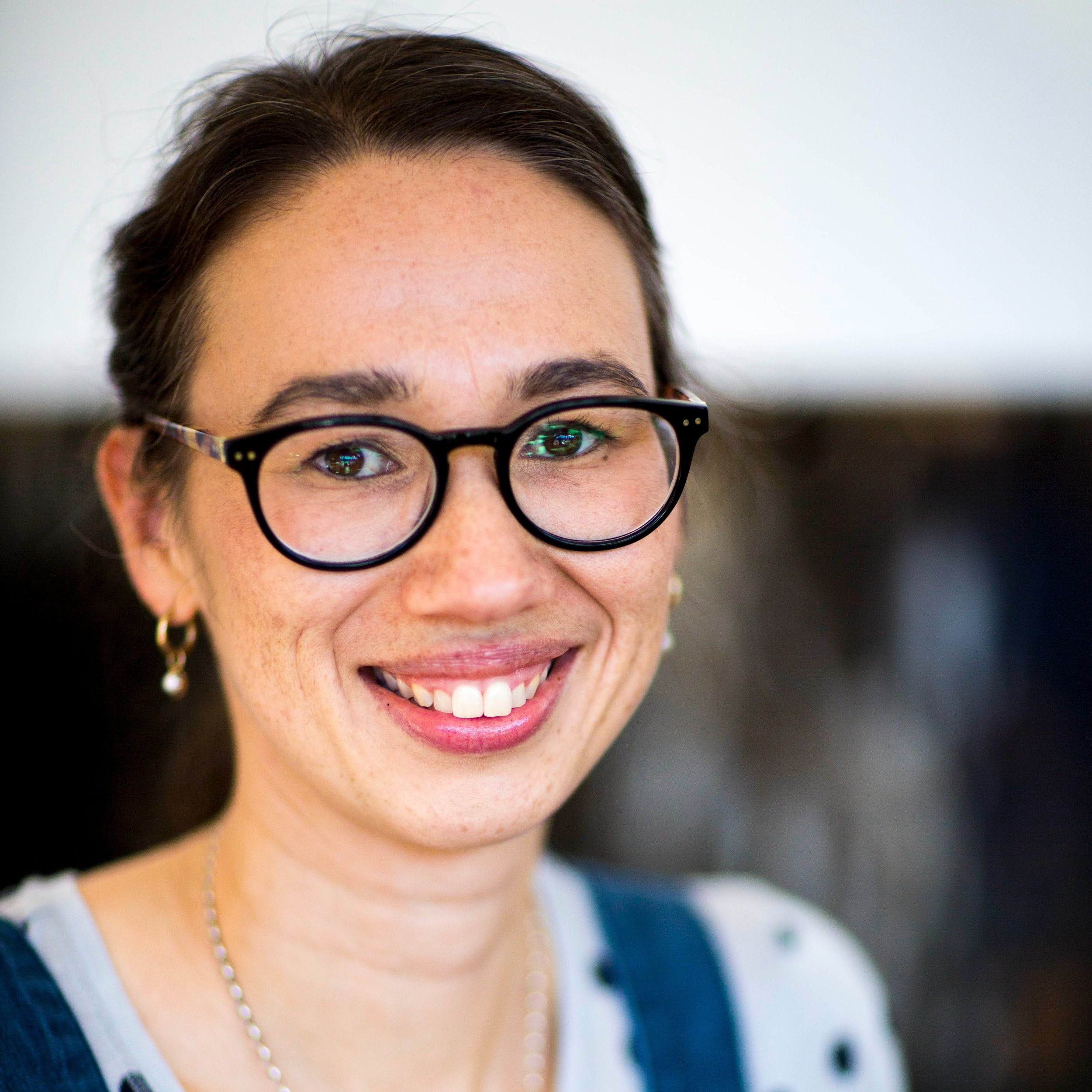 A woman smiling, showing her teeth, wearing black glasses, a pearl necklace, and earrings, with a white background.