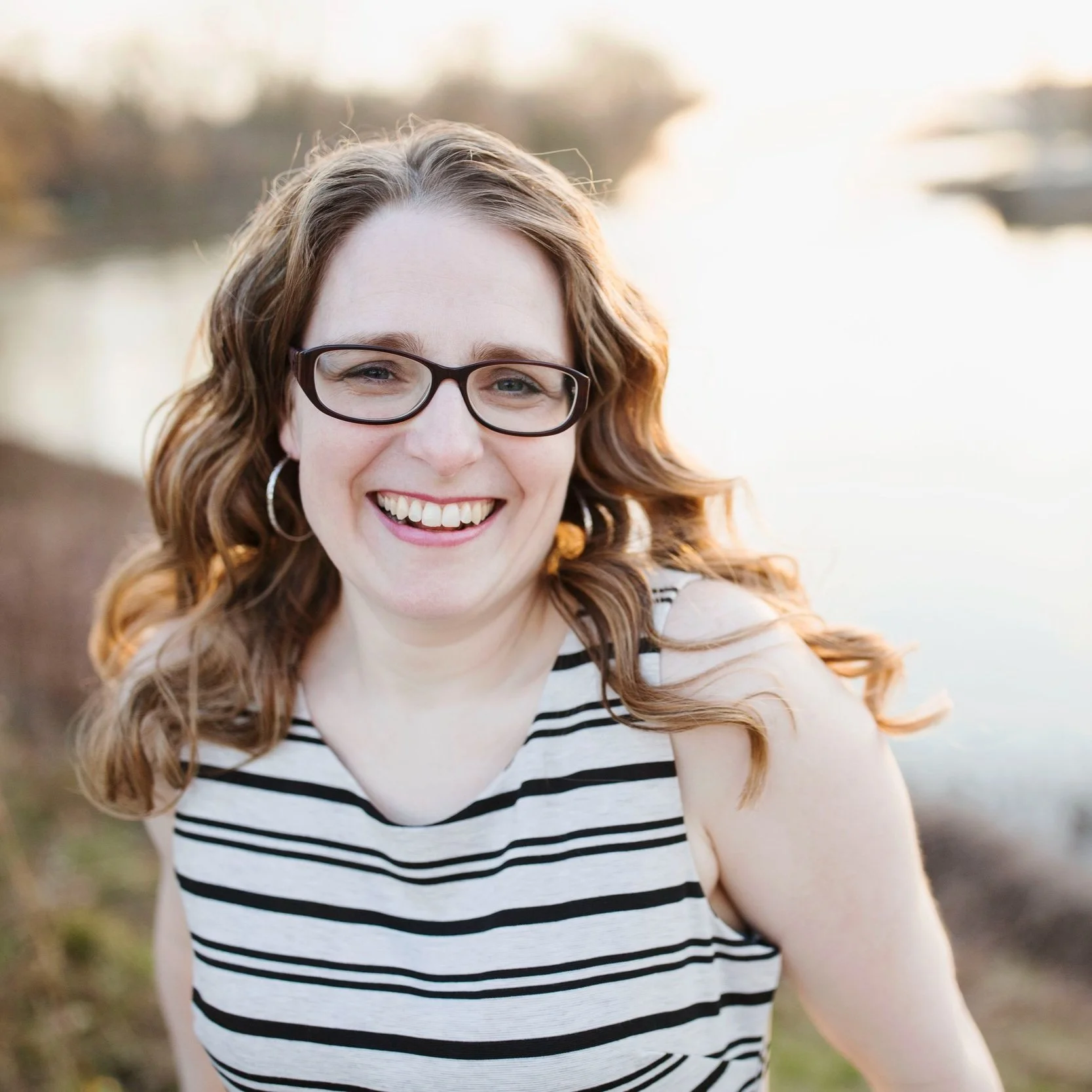 A woman with red hair, glasses, and hoop earrings smiling outdoors during sunset, wearing a black and white striped sleeveless top.