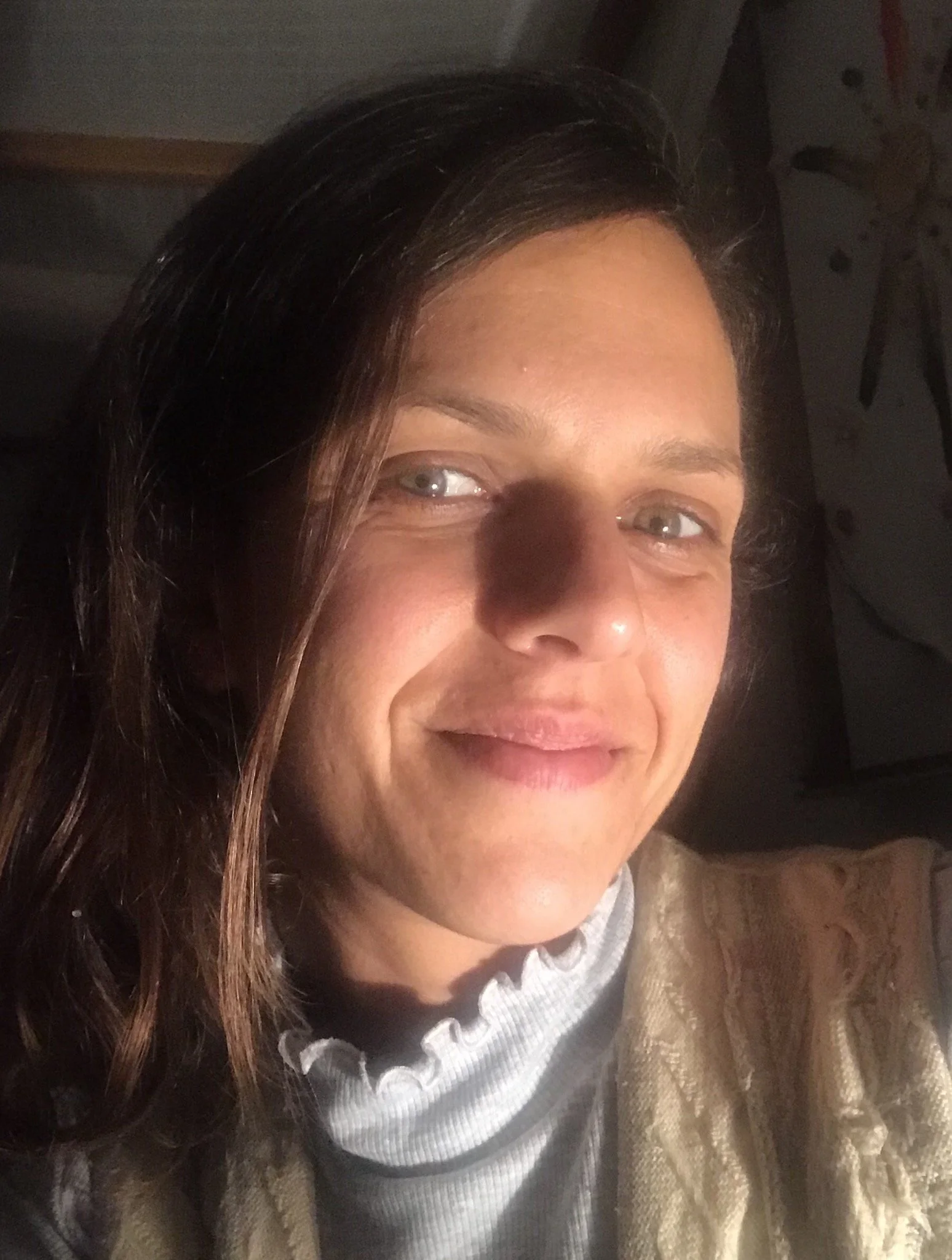 A woman with long brown hair and light eyes smiling, wearing a gold-colored jacket and a white ruffled blouse, sitting indoors with a dark background.