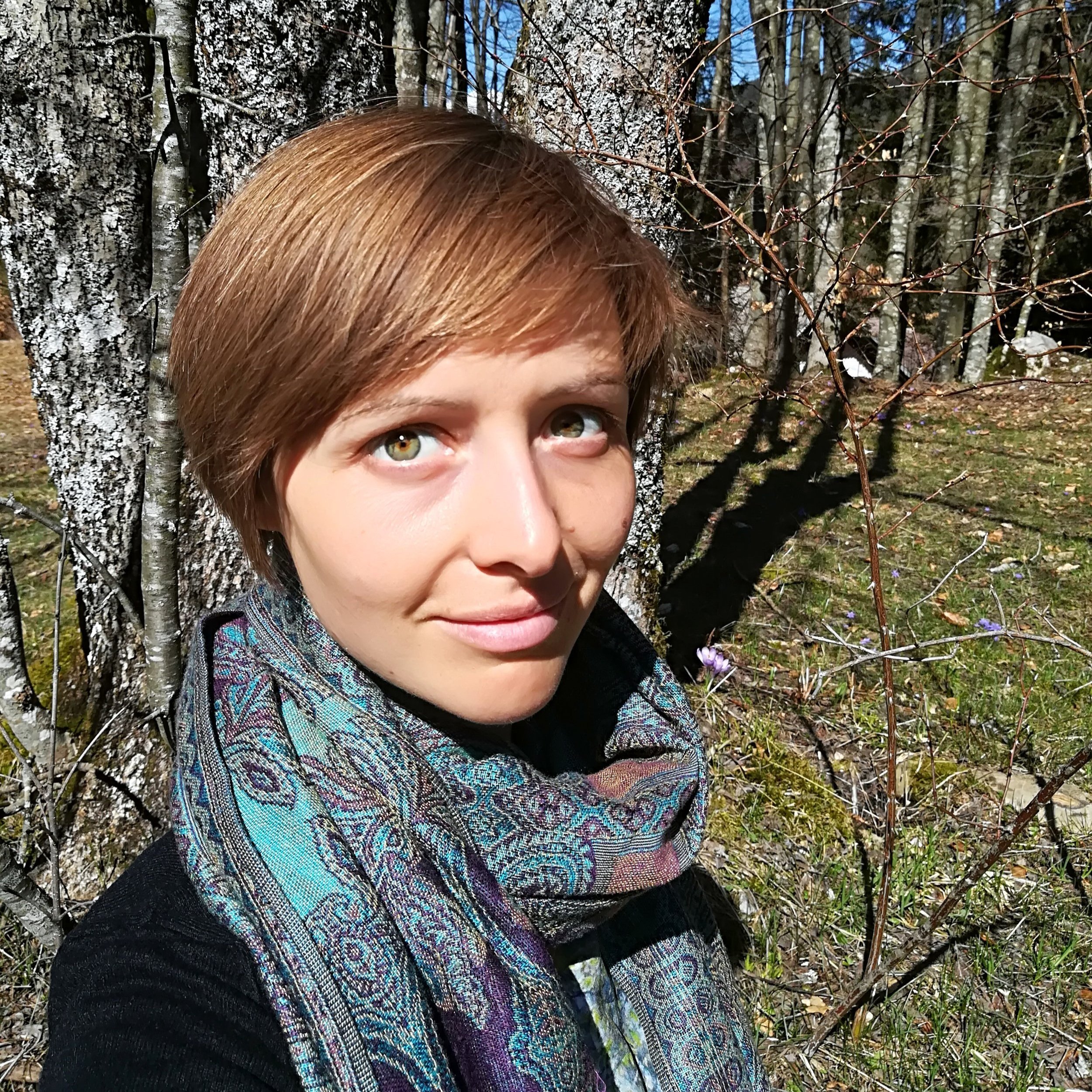Close-up of a person with short reddish-brown hair and fair skin, outdoors in a forest with trees covered in white bark and some purple flowers on the ground.