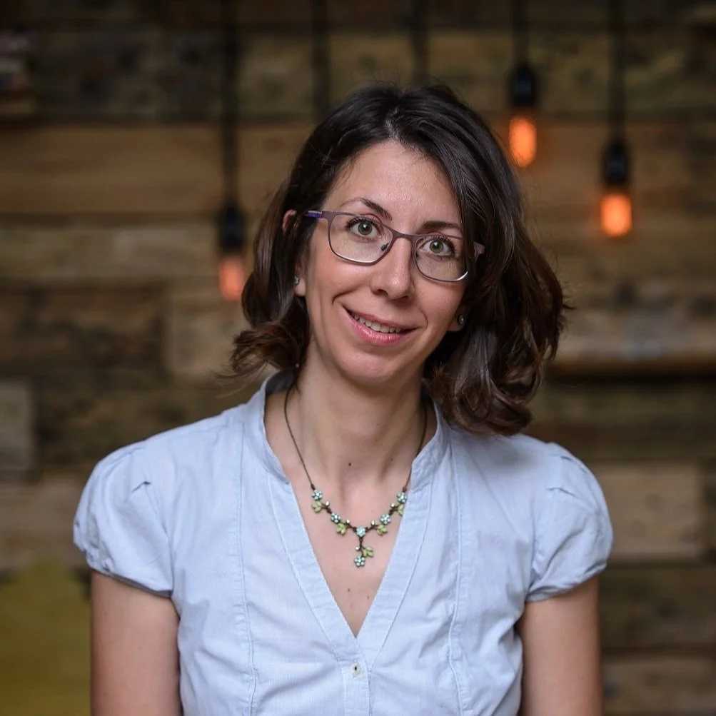A woman with glasses and dark brown hair smiling, wearing a light blue blouse and a floral necklace, standing in front of a wooden wall with hanging Edison style light bulbs.
