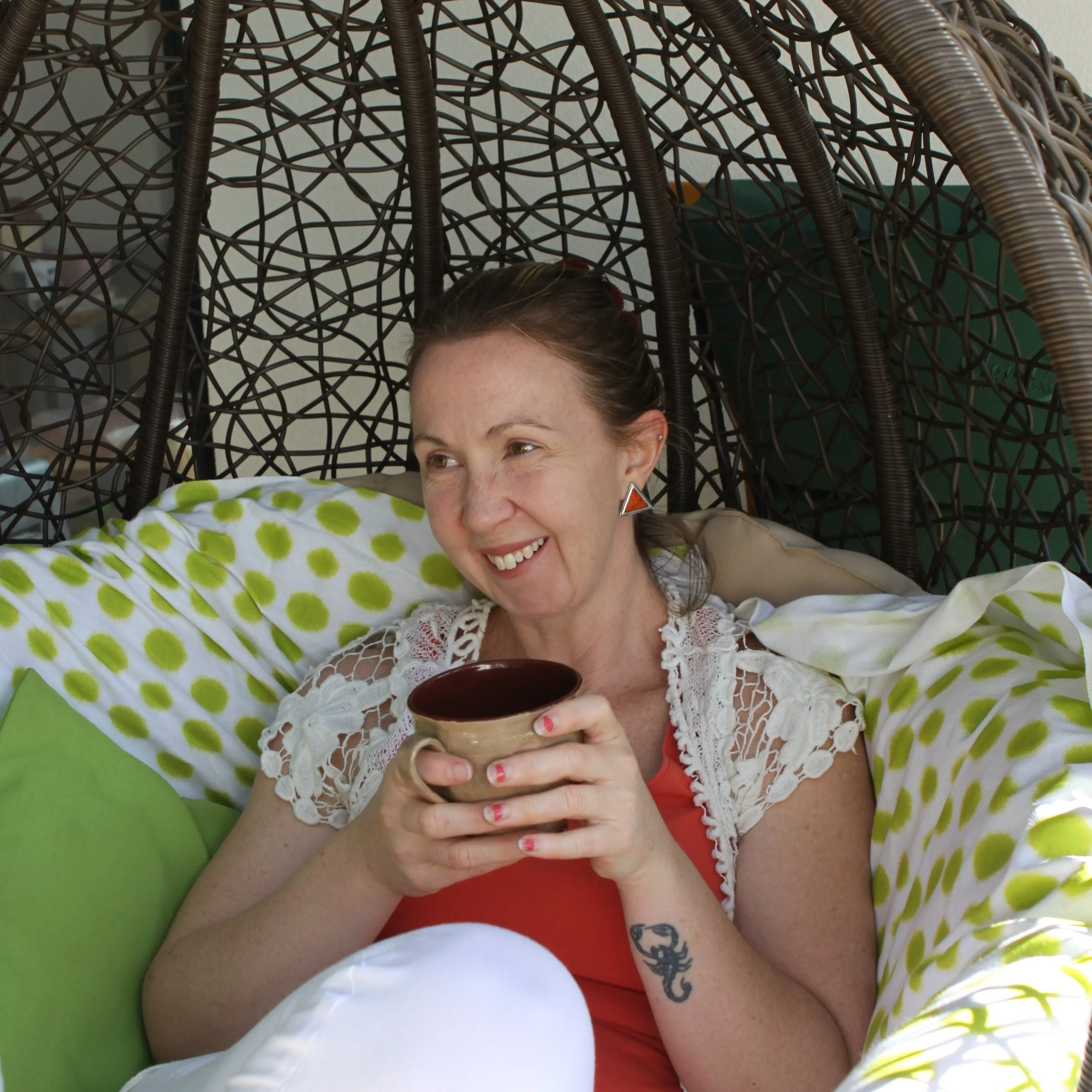 A woman with short brown hair, smiling, holding a cup of coffee, lying on a green polka dot blanket with a wicker hanging chair overhead, part of her tattooed arm is visible.