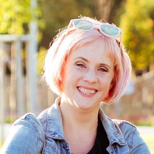 Young woman with short pink hair wearing sunglasses on her head, smiling outdoors during daytime.
