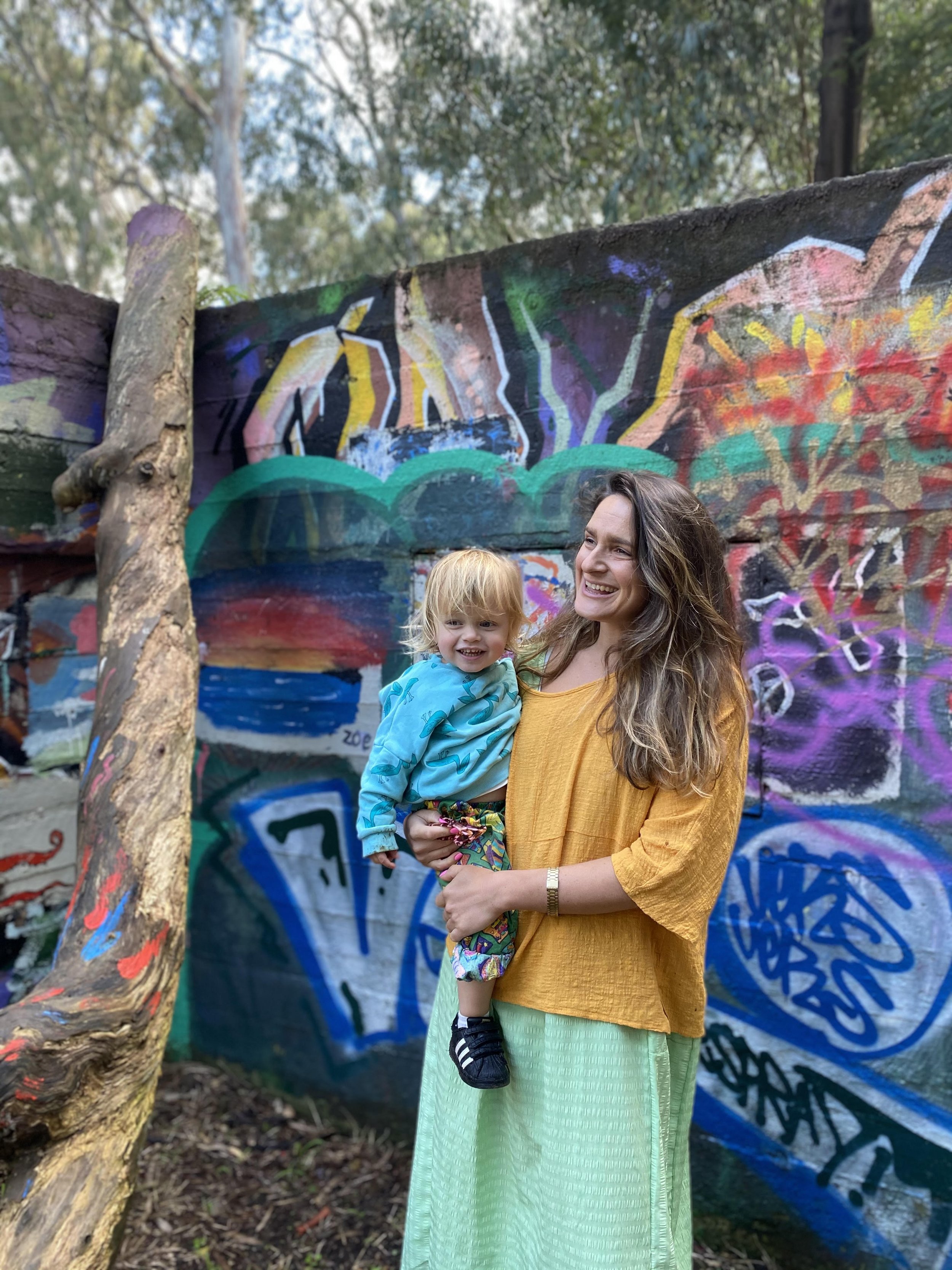 A woman with long wavy brown hair wearing a yellow top and green skirt is holding a young blonde girl in a blue patterned jacket and colorful pants in front of a graffiti-covered wall in a wooded area.
