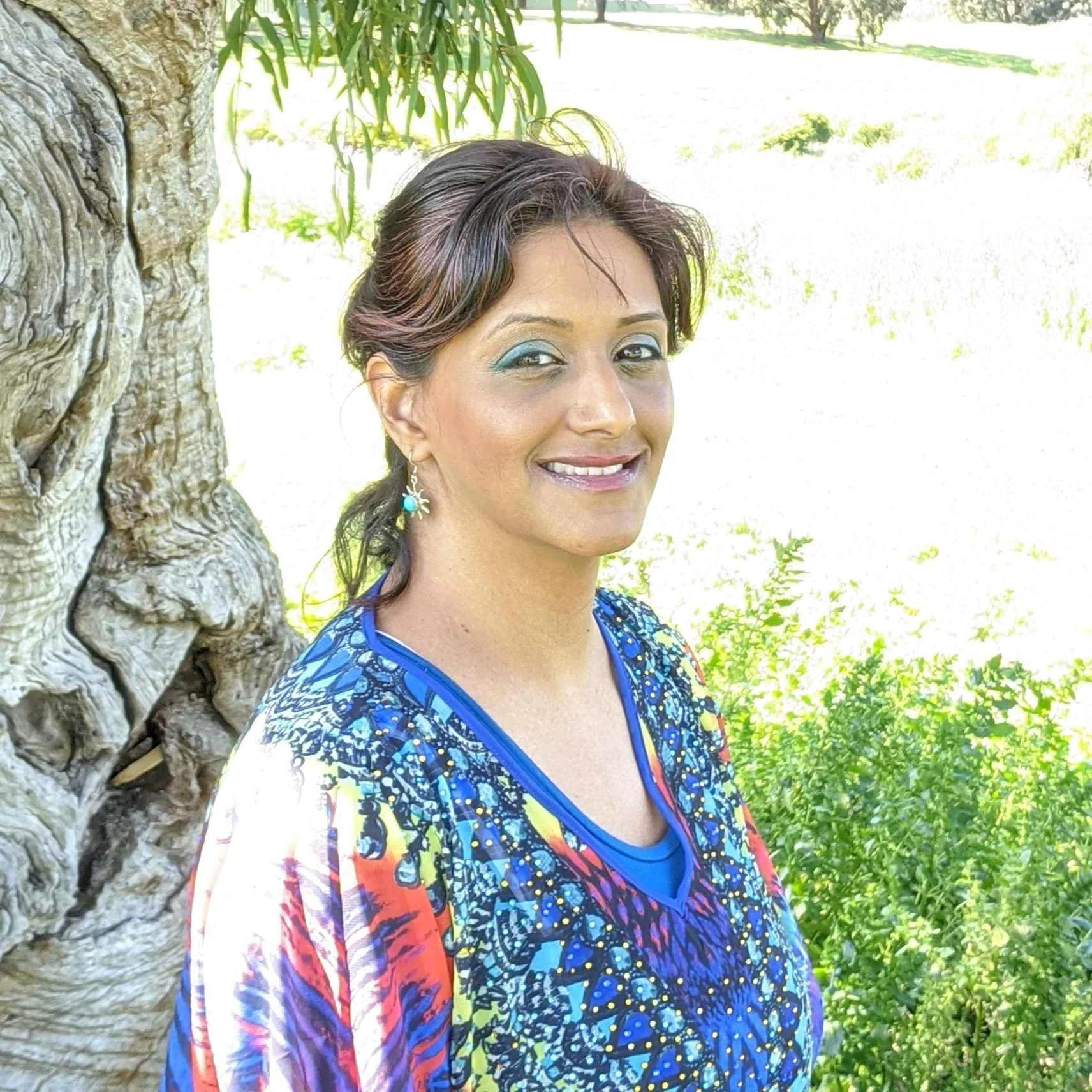 A woman with short, dark hair and turquoise makeup around her eyes, smiling, standing next to a large tree with textured bark in a sunny outdoor setting with green foliage in the background.