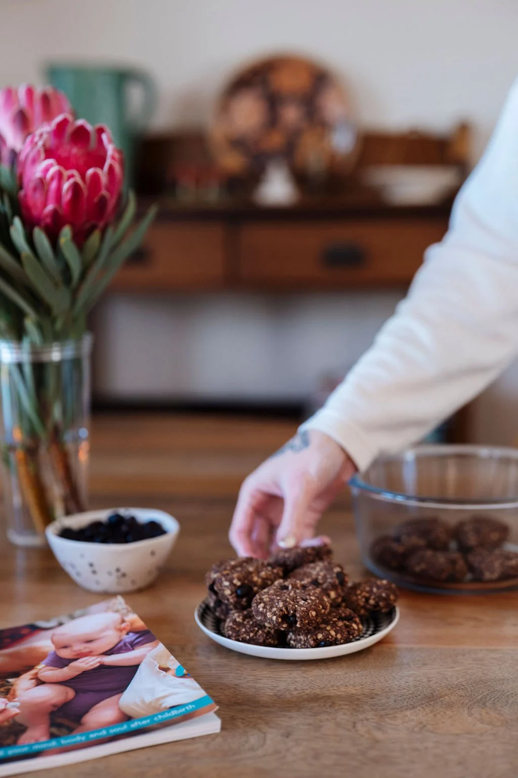 A certified doula reaching for homemade chocolate cookies on a white plate on a wooden table, with a vase of pink protea flowers, a bowl of blackberries, and a cookbook in the background.