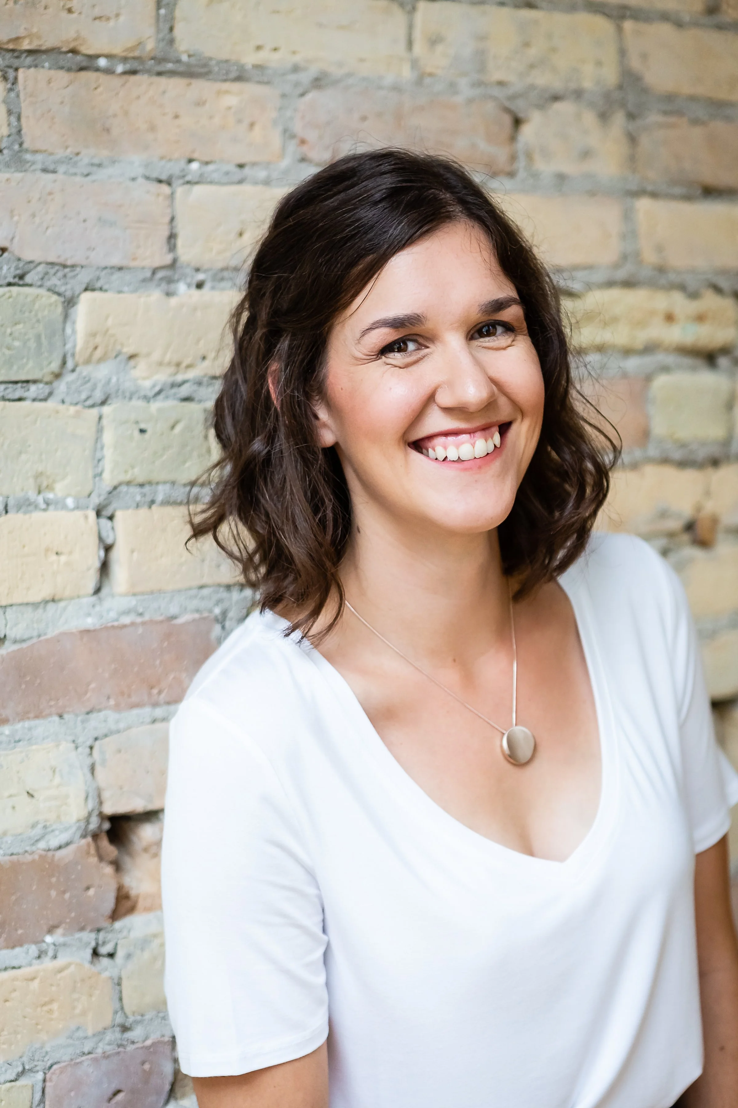 A smiling woman with wavy brown hair wearing a white t-shirt and a round pendant necklace, standing in front of a brick wall.