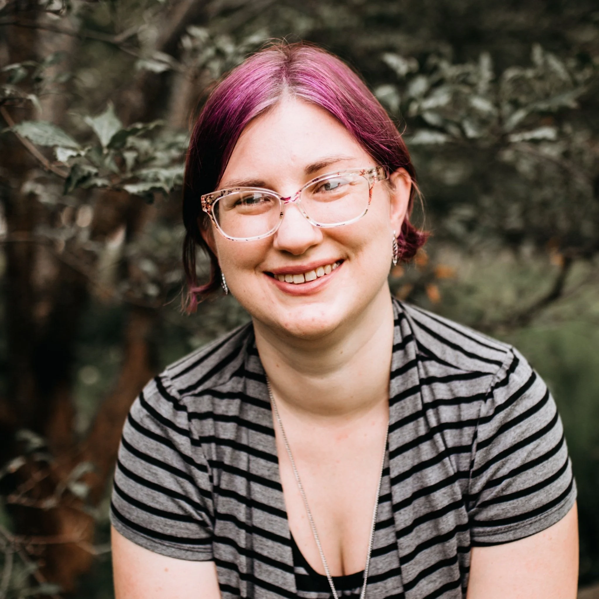 Person with short hair dyed pink and purple, wearing clear glasses with tortoiseshell pattern, smiling, in outdoor setting with leafy background, wearing a black and white striped top.