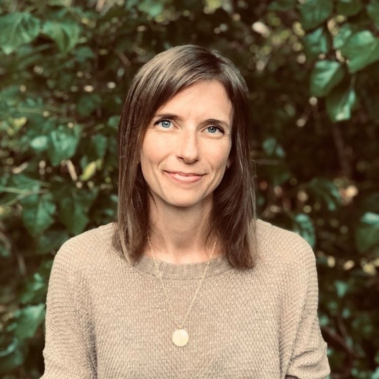 A woman with shoulder-length brown hair and blue eyes, smiling softly, wearing a beige knit sweater and a pendant necklace, with a background of green foliage.