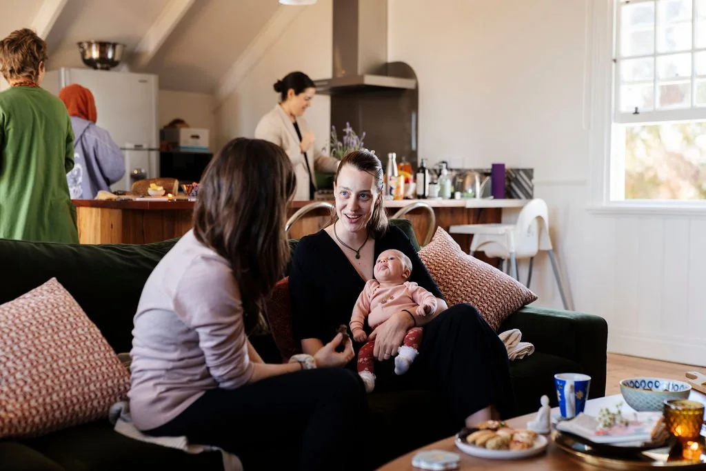 Two women sitting on a sofa engaged in conversation, one holding a baby, with a busy kitchen in the background, during a casual gathering in a home.