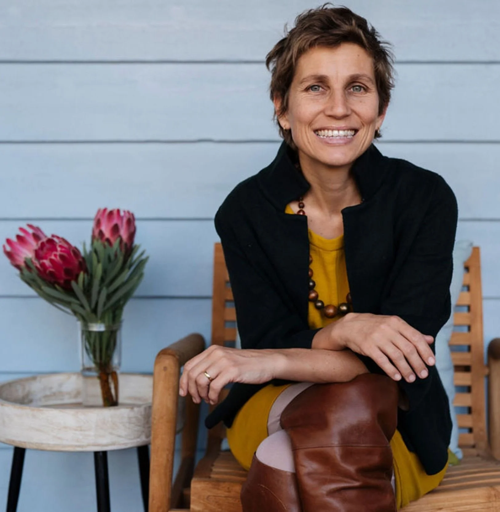 A smiling woman with short brown hair and blue eyes sitting at a wooden table, wearing a yellow dress, black jacket, and a beaded necklace, with a vase of pink protea flowers and a wooden chair with a slatted backrest behind her.