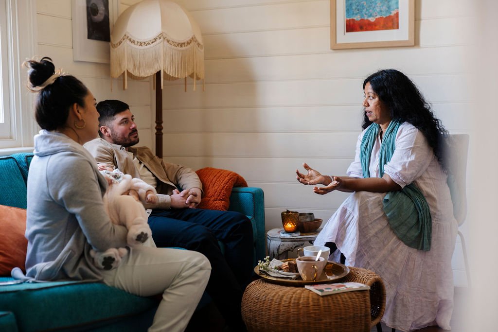 A dark skinned woman sits across from a man and woman holding a baby. She holds her palms out and is listening intently