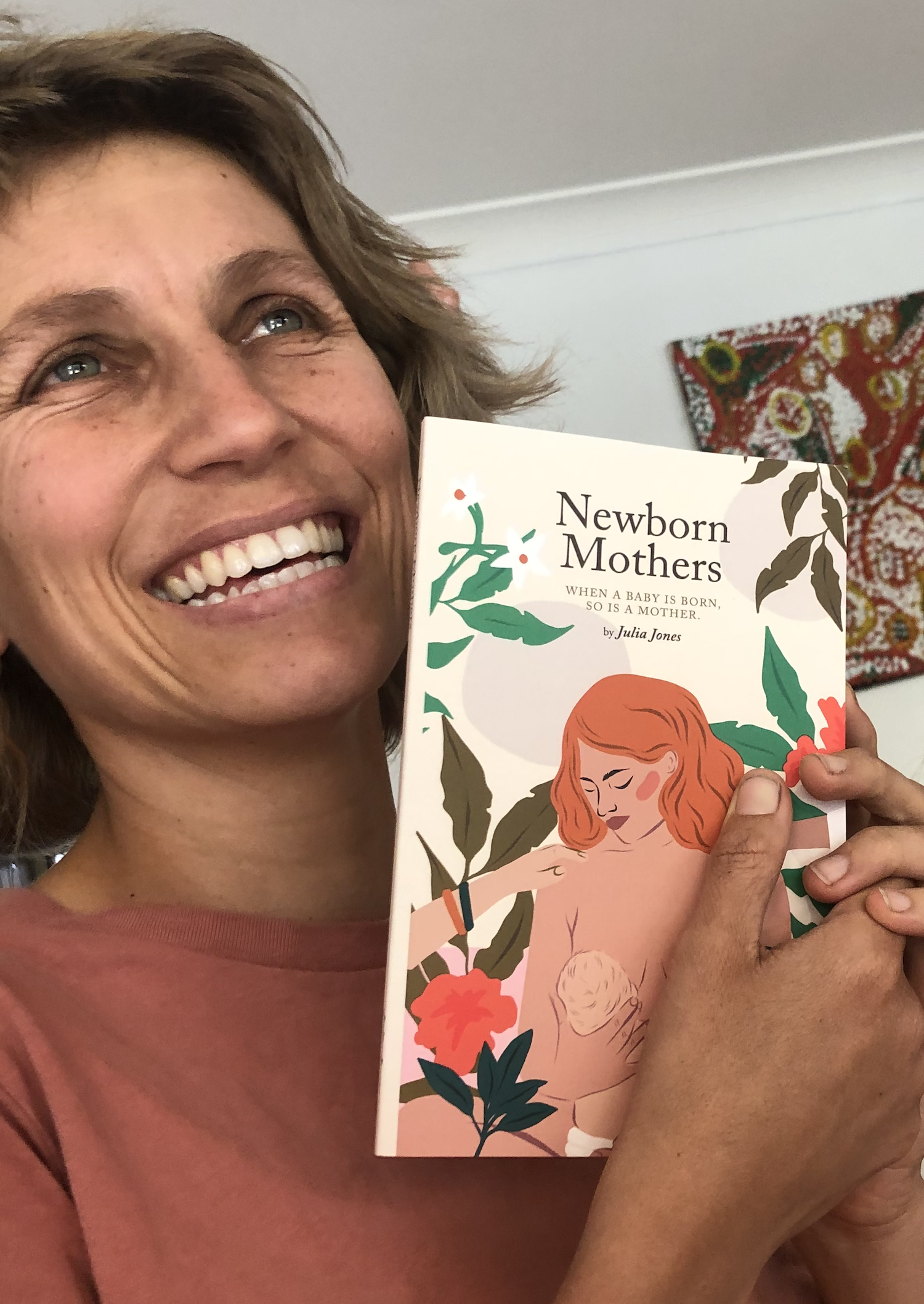 Julia Jones holding the first printed copy of her book titled 'Newborn Mothers' featuring an illustration of a breastfeeding woman with red hair and flowers in her hand.