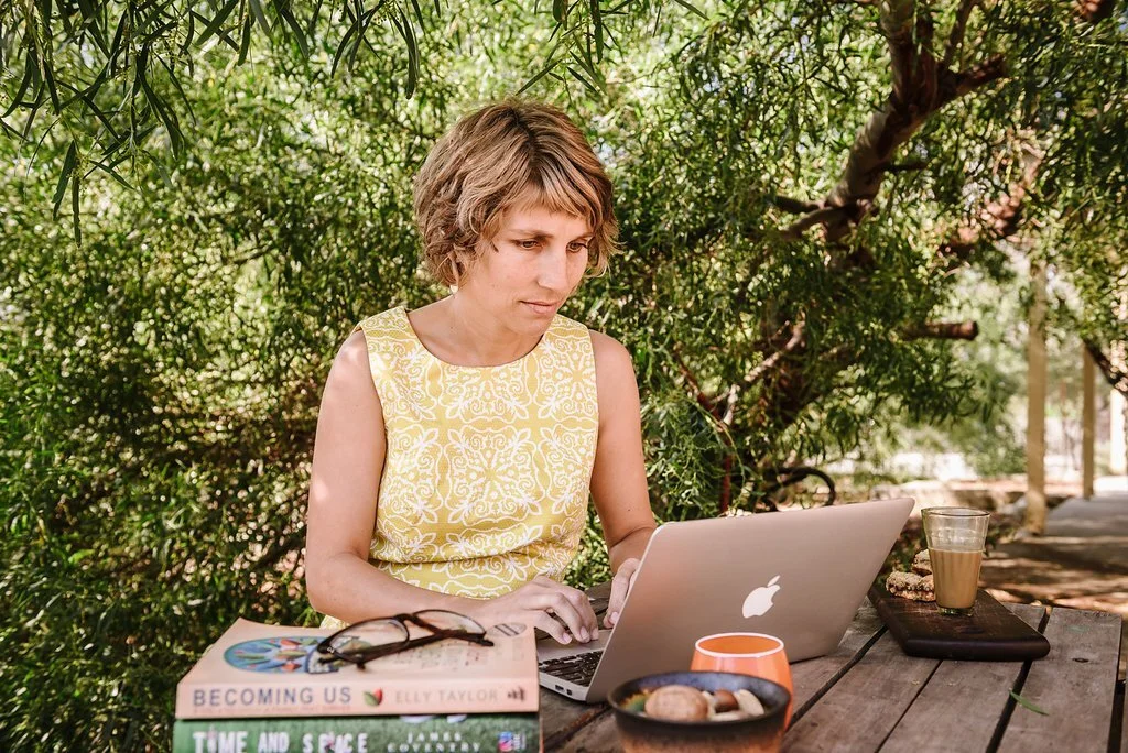 Woman in yellow patterned sleeveless top working on a silver MacBook at an outdoor wooden table surrounded by greenery.