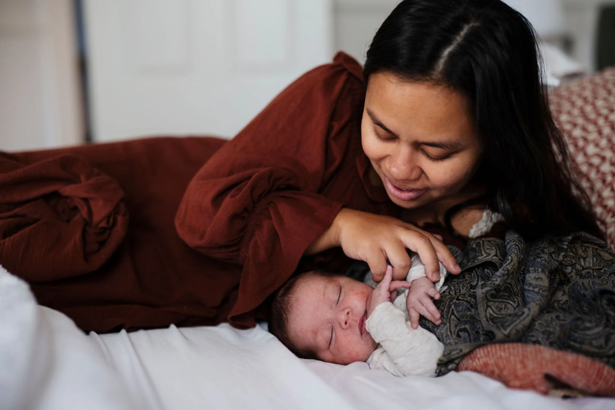 A woman with black hair and a reddish-brown shirt is lying on a bed, gently cradling a newborn baby with a woman lying underneath. The scene appears intimate and tender.