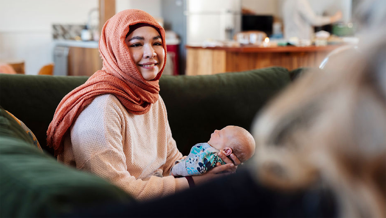 A woman in a peach-colored sweater and a rust-colored hijab sitting on a green sofa, holding a newborn baby wrapped in a patterned blanket, smiling at the camera behind a dog in the foreground.