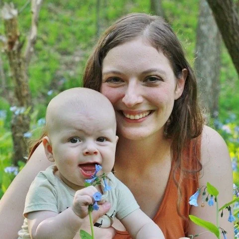 A woman smiling with a baby in a garden with green foliage and blue flowers.