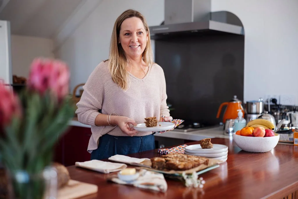 A postpartum doula in a kitchen holding a plate with a piece of cake, smiling. The kitchen counter has a bowl of fruit, a cake, and various kitchen items.