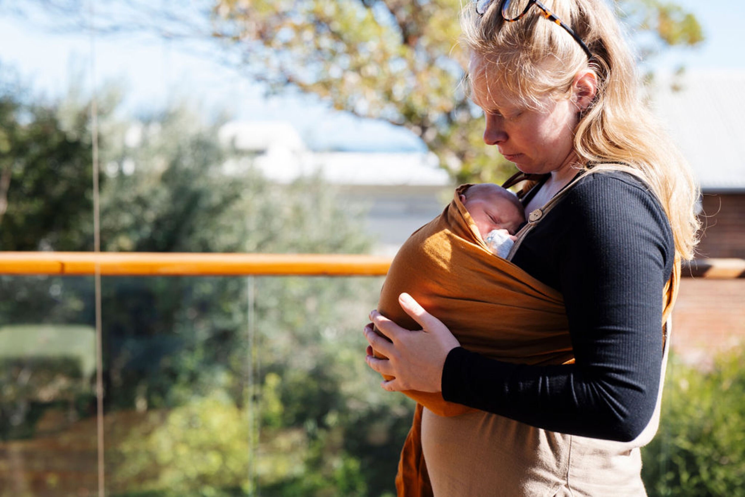Postpartum mother looking down at her sleeping newborn in a brown baby carrier outdoors