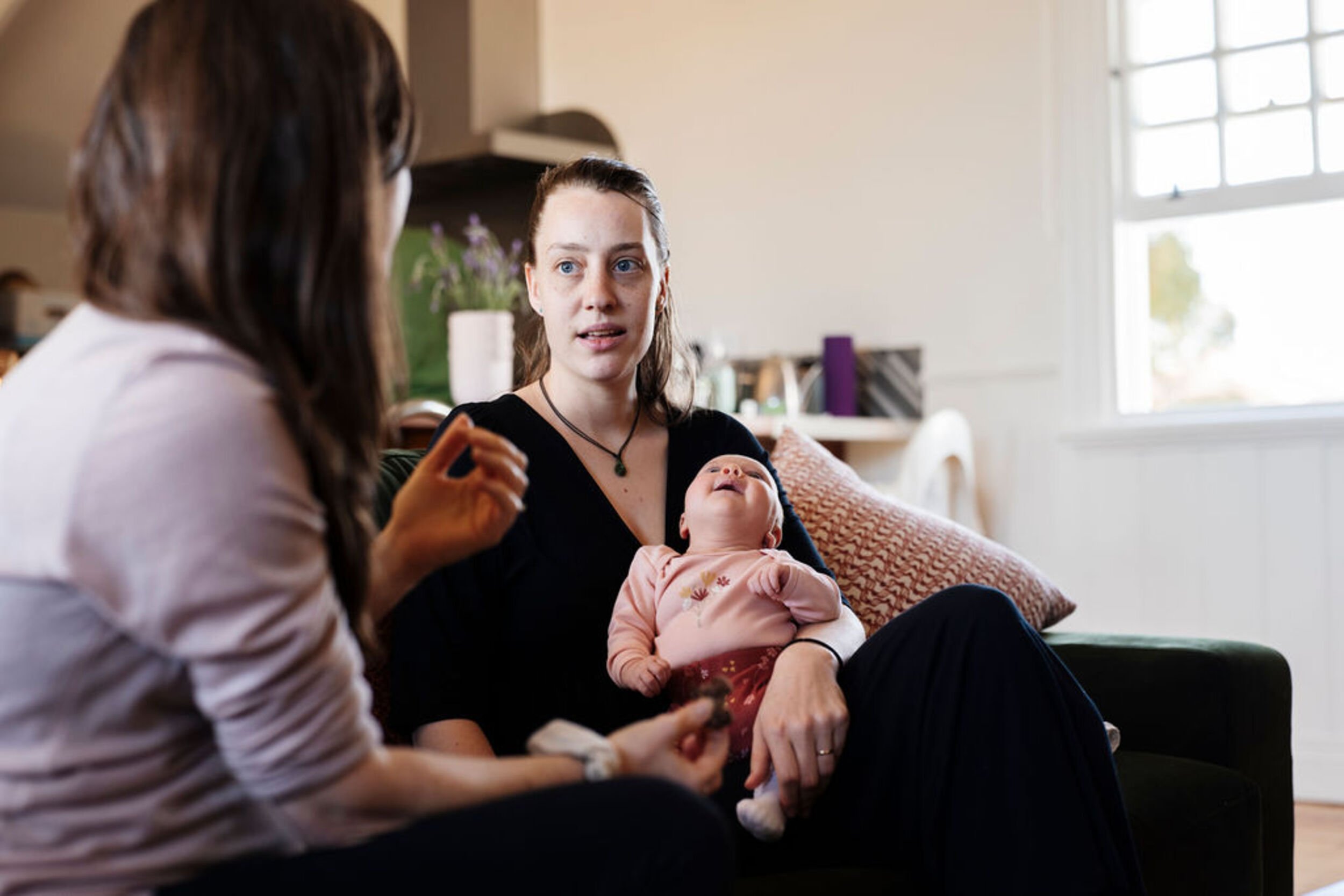 A mother holding a baby and a postpartum doula are seated and engaging in conversation in a bright, cozy living room.