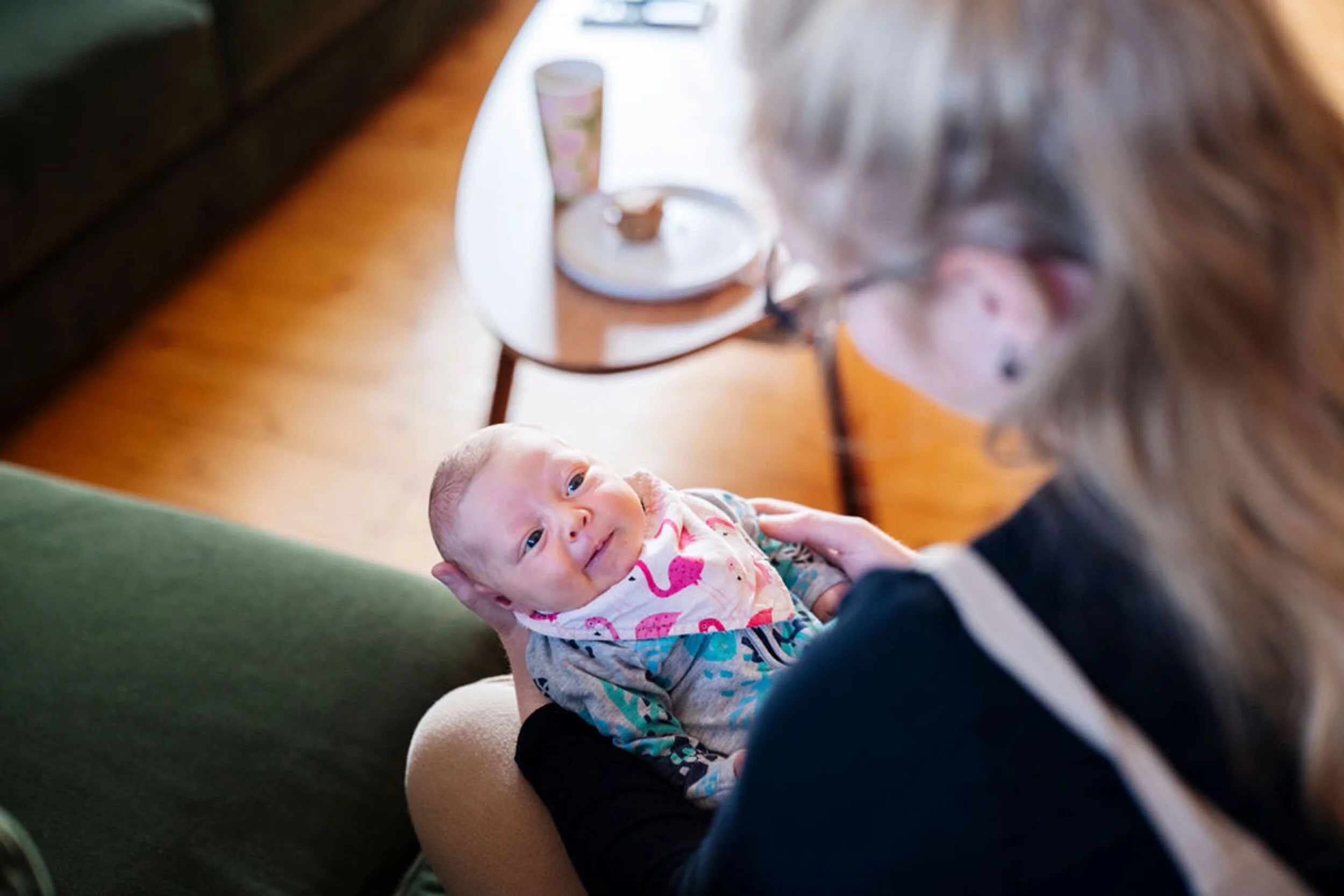 A baby lying on a mother's lap, looking up with a smile, while the mother wearing glasses interacts with the baby.